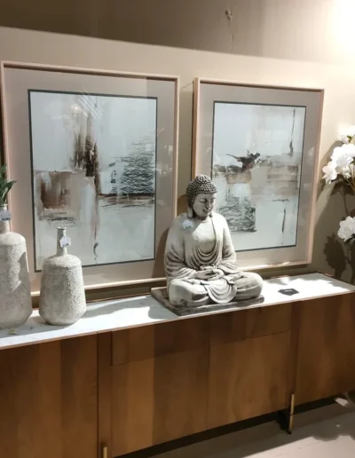 Mueble aparador de madera con encimera blanca, decorado con una escultura de Buda, jarrones texturizados y dos cuadros grandes de arte abstracto en tonos neutros.