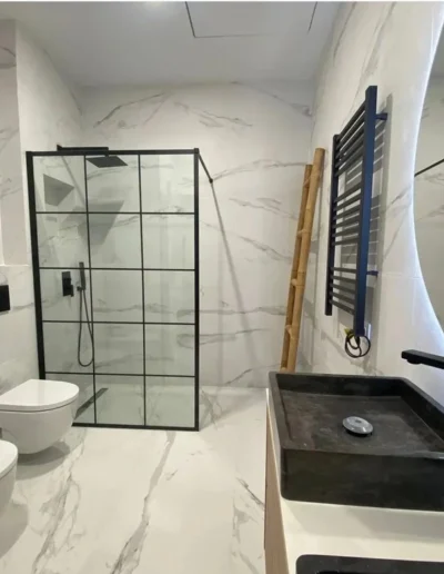 Reforma de baño moderno con azulejos de efecto mármol blanco, mampara de ducha con perfilería negra cuadriculada, sanitarios suspendidos y lavabo sobre encimera oscuro