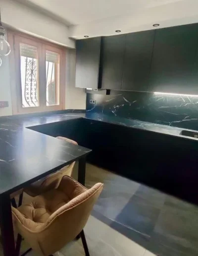 Diseño de cocina moderna en color negro mate con encimera y salpicadero de efecto mármol oscuro con vetas blancas, iluminación led bajo los muebles altos y una mesa a juego con sillas tapizadas.