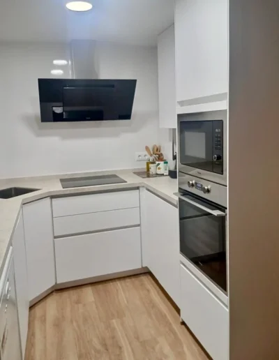 Reforma de cocina pequeña y moderna distribuida en forma de U, con muebles blancos sin tiradores, columna de horno y microondas, campana extractora decorativa negra y suelo laminado de madera clara.