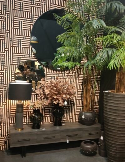 Decoración de recibidor o salón con mueble bajo de madera oscura, un gran espejo redondo negro, lámpara de mesa, jarrones y plantas tropicales sobre un fondo de papel pintado geométrico.
