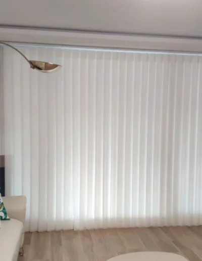Gran ventanal de salón decorado con cortinas de onda perfecta en color blanco translúcido, caídas de cortina laterales tricolor, sofá beige y una lámpara de pie arqueada dorada.