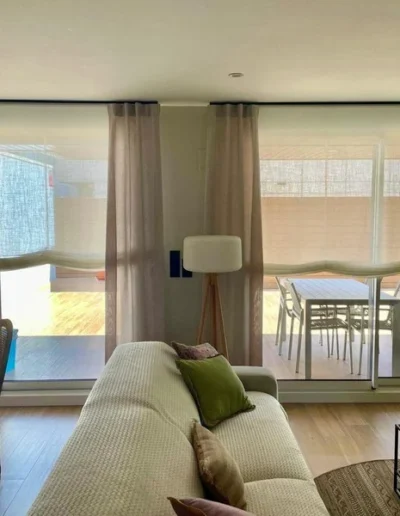 Decoración de salón comedor luminoso con sofá beige, cojines verdes, mesa de comedor redonda y grandes ventanales vestidos con estores paqueto y cortinas que dan acceso a una terraza con mesa exterior.